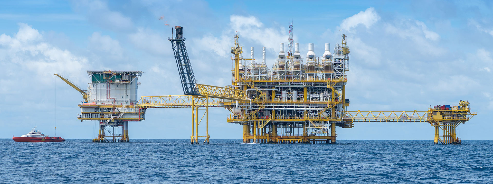 The type of offshore oil rig that might use BarCan barite API Drilling mud. Rig has no identifying features – its yellow structure is shown on a bright, sunny day in the middle of the ocean. A platform supply vessel in the picture gives a sense of how immense the oil rig is.